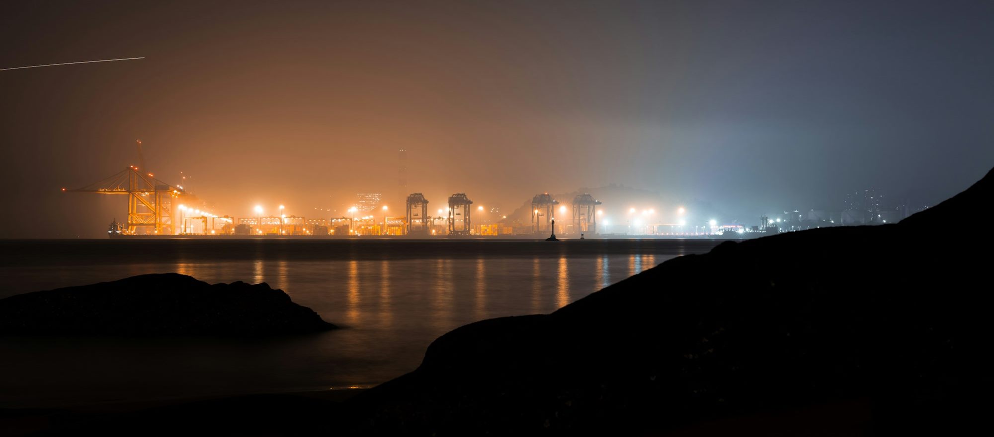 A container port lit at night across the water, its cranes and lights reflected in the sea