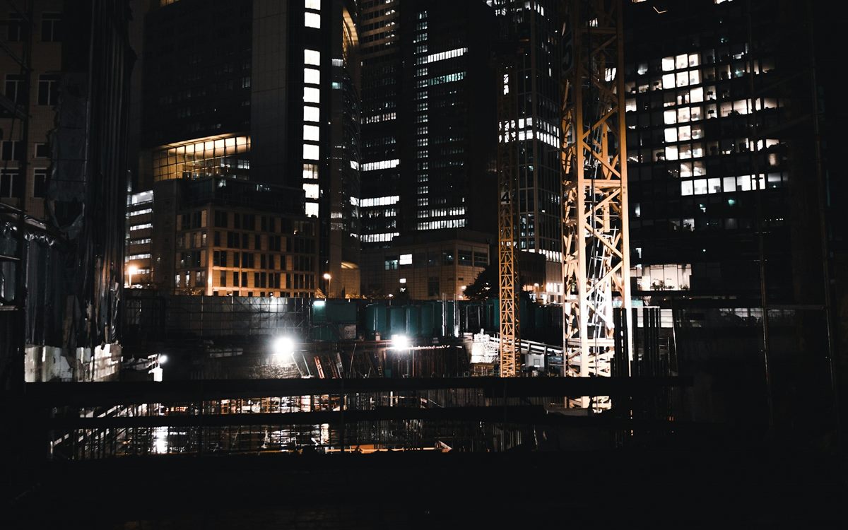 A construction pit lit by floodlights at night among high-rise buildings