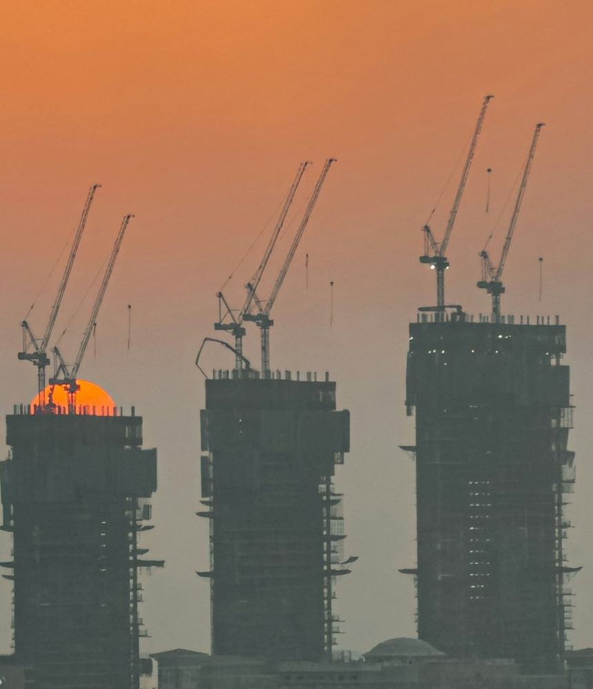 Three towers under construction topped by cranes at sunset